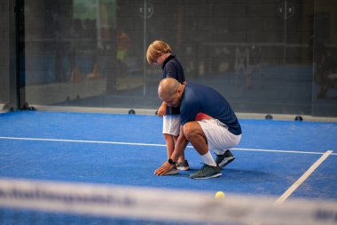 Öğrencisi Padel 'den çocuğa ders verirken gözlemci, küçük çocuğa kapalı tenis kortunda nasıl padel oynanacağını öğretiyor.