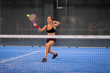 Kapalı alanda padel oynayan bir kadın genç sporcu kadın raketle topa vuruyor.