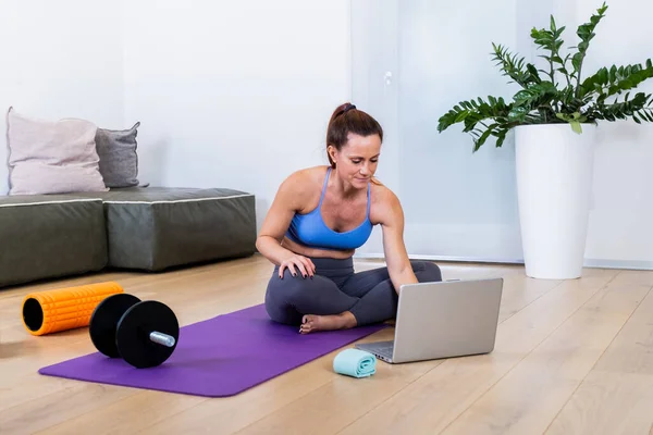 Fit young woman in sports clothes searching for training video course at web using laptop, lying on floor yoga mat in the modern living room with gym equipment