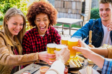 Group of multiethnic friends living healthy lifestyle and smiling and joking while drinking beer at outdoor pub restaurant - Young people toasting with beer glass during happy hour at bar