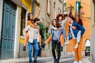Group of friends having fun walking on city street at holiday - Happy young multi-ethnic men giving their girlfriends piggyback ride