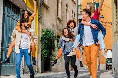 Group of friends having fun walking on city street at holiday - Happy young multi-ethnic men giving their girlfriends piggyback ride