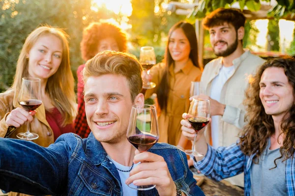 Group of multiracial friends taking selfies and celebrating with wine glasses at dinner time in terrace - Young people eating and drinking red wine at the outdoor rooftop bar  and taking selfie using mobile smartphone