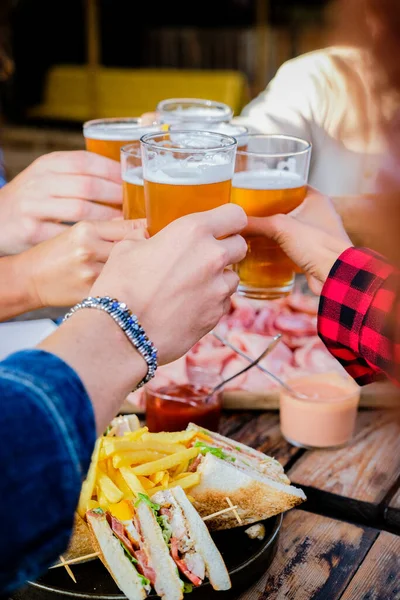 Close up of hands of multiethnic drinking beer at outdoor pub restaurant - Young people enjoying drinks during happy hour at terrace bar toasting with beers - Friendship concept