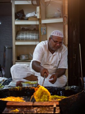 Mumbai, India - May 1, 2022 : Muslim male vendor cooking selling halal foods and snacks fried pancake Malpuas from roadside stall at night market in holy month of Ramadan. Vertical or portrait orientation