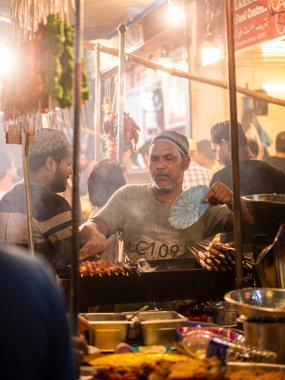 Mumbai, India - May 1, 2022 : Muslim male vendor cooking selling halal foods and Kababs from roadside stall at night market in holy month of Ramadan . Vertical or portrait orientation