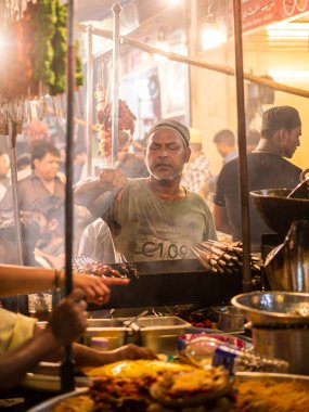 Mumbai, India - May 1, 2022 : Muslim male vendor cooking selling halal foods and Kababs from roadside stall at night market in holy month of Ramadan . Vertical or portrait orientation