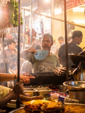 Mumbai, India - May 1, 2022 : Muslim male vendor cooking selling halal foods and Kababs from roadside stall at night market in holy month of Ramadan . Vertical or portrait orientation