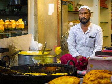 Mumbai, India - May 1, 2022 : Muslim male vendor cooking selling halal foods and snacks fried pancake Malpuas from roadside stall at night market in holy month of Ramadan Ramazan at Bhendi Bazar