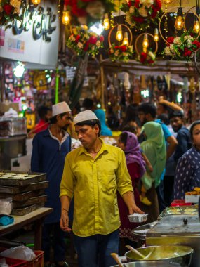 Mumbai, India - May 1, 2022 : Muslim male vendor cooking selling halal foods and varous sweets from roadside stall at night market in holy month of Ramadan . Vertical or portrait orientation