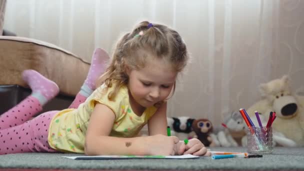 En liten flicka ritar med filtspets pennor på papper, liggande på golvet hemma. Konst, utbildning, barndom, begrepp. — Stockvideo