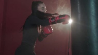 Genç güçlü esmer kadın güçlü bir şekilde kum torbasına vuruyor ve tekmeliyor. Kız boks salonunda yarışmalara hazırlanıyor. Koyu renk bir spor salonunda güzel bir kadın kickboksçunun eğitimi. Yavaş çekim.