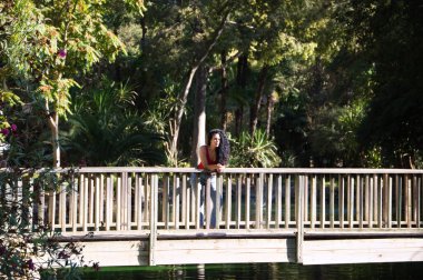 Güzel, yetişkin bir kadın, kıvırcık saçlı ve Latin kökenli esmer, parktaki bir gölün ahşap korkuluğuna yaslanmış kameraya bakarken farklı ifadeler takınıyor. Tatil ve seyahat kavramı.