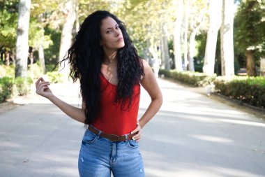 Güzel, yetişkin bir kadın, kıvırcık saçlı ve çingene latincesi özellikli esmer bir yolda ve kenarlarında dev ağaçlar olan kadın farklı jestler yapıyor ve poz veriyor. İfadeleri kavra