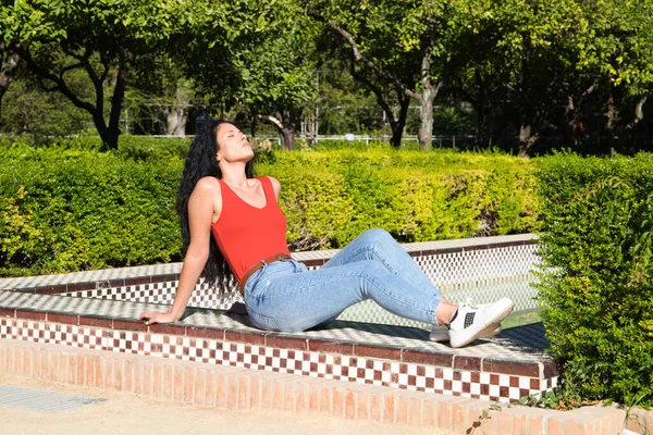 ラテン語の特徴を持つ美しい 大人の 暗い髪の 巻き毛の女性は 公園のタイル張りのベンチで日光浴です 四季のコンセプト — ストック写真