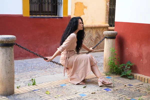 在典型的地中海风格的城市街道上 一位美丽的女人坐在链条上 一头乌黑的卷曲的头发 这个女人是严肃而悲伤的 概念表述 — 图库照片