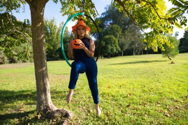 Parkta spor yapan yetişkin kadın bir akrobatik çember atleti. Kadının elinde turuncu bir cadılar bayramı şapkası ve akrobasi yaparken bir balkabağı var. Sağlık ve spor konsepti.