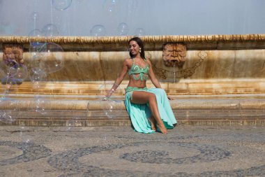 Genç ve güzel bir dansçı büyük bir meydanda bir çeşmenin kenarında oturuyor. O yalınayak ve saçları gevşek. O, mavi bir elbise giyiyor. Moda ve güzellik konsepti