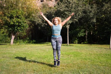 Parkta spor yapan güzel, şişman sarışın kadın. Çimlerin üzerinde zıplıyor, kollarını ve bacaklarını açıyor. Açık hava sporu ve jimnastik konsepti. Sağlıklı yaşam.