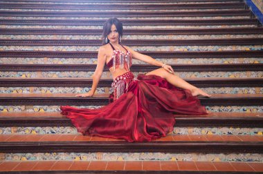 Genç ve güzel dansçı bir merdivende oturmuş dans mankeni pozları veriyor. Dansçı kırmızı bir elbise giyiyor. Göbek dansı konsepti.