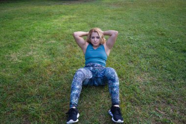 Güzel sarışın kadın çimlerin üzerindeki parkta jimnastik ve karın egzersizi yapıyor. Kadın şişman ve spor yapması ve sağlıklı bir diyet yapması gerekiyor. Jimnastik konsepti ve sağlıklı beslenme.