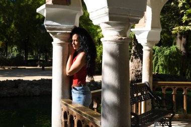 Koyu renk saçlı, kıvırcık saçlı, parktaki çardağın sütunlarına yaslanmış Latin bir kadın. Kadın üzgün, çünkü o ortağını bekliyor ve o gelmiyor. Kadının acelesi var.