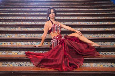Genç ve güzel dansçı bir merdivende oturmuş dans mankeni pozları veriyor. Dansçı kırmızı bir elbise giyiyor. Göbek dansı konsepti.