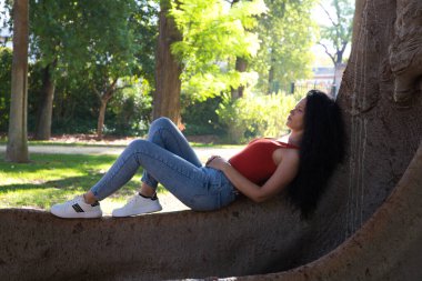 Güzel, yetişkin, siyah saçlı, kıvırcık saçlı, Latin çingenenin özelliklerine sahip bir kadın büyük bir şehir parkındaki dev bir incir ağacının köklerinde yatıyor. Rahatlayın ve tatil konsepti.