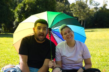 İkili cinsiyet ayrımı yapmayan bir insan ve eşcinsel bir adam parkta oturmuş, kendilerini güneşten korurlar. Kameraya poz veriyorlar ve çok mutlular. Çeşitlilik ve gay gururu.