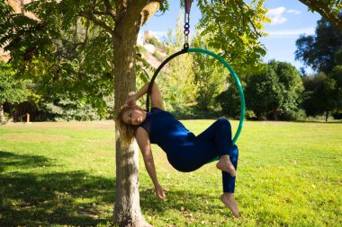 Sarışın kadın ve genç jimnastikçi akrobat sporcu parkta hava halkası üzerinde egzersiz yapıyorlar. Mavi kostümlü kıvrak kadın sirk sanatçılarının kalçalarıyla dans edişini canlandırıyor..