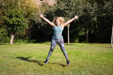 Parkta spor yapan güzel, şişman sarışın kadın. Çimlerin üzerinde zıplıyor, kollarını ve bacaklarını açıyor. Açık hava sporu ve jimnastik konsepti. Sağlıklı yaşam.
