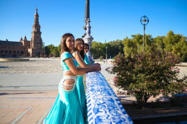 Açık mavi giyimli üç dansçı bir köprünün korkuluklarına yaslanıyorlar. Birbirleriyle konuşuyorlar ve kameraya bakıyorlar. Göbek dansı ve Hint dansı konsepti. Folklor