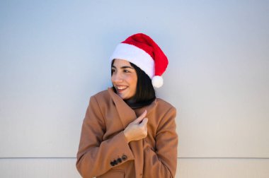 Genç, güzel, koyu renk saçlı kadın kendini soğuktan korumak için Noel Baba şapkası ve kahverengi bir ceket giyiyor. Kadın farklı pozlar veriyor. Mutlu noeller ve mutlu yıllar..