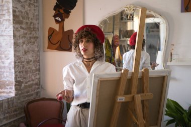 Genç, güzel, girişken bir kadın. Resim ve moda tasarımı sanatına adanmış sanatçı, kafasına kırmızı bir bere takıyor ve sehpanın üzerindeki tuvale resim yapıyor. Girişimci kadın konsepti