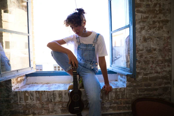 Genç ve güzel bir kadın stüdyosunda oturmuş ukulele çalıyor ve müzikten esinlenerek pencereden dışarı bakıyor. Müzik ve sanat konsepti.