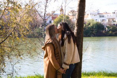 Parkta ağaca yaslanan lezbiyen çift ellerini birbirine kenetledi. Bakışlar ve kucaklaşmalarla etkileşime girerken çok mutlu ve aşık görünüyorlar. Homoseksüellik kavramı, lezbiyenler.