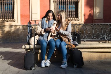 Lezbiyen çift kentteki devasa bir meydanda bankta oturuyor. Tatilde şehre geldiklerinde cep telefonlarına danışıyorlar ve bavulları yanlarında. Turizm.