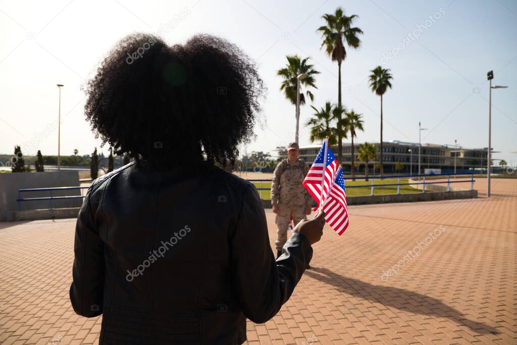 Young Afro-American woman holds two American flags in her hand while waiting for her boyfriend who has come home from the war. An American soldier can be seen in the background. Concept of war.
