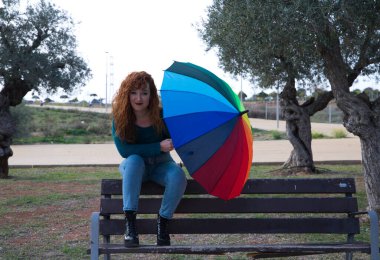 Genç ve güzel kızıl saçlı kadın birçok zeytin ağacının olduğu bir parkta bir bankta oturuyor. Gri günü aydınlatmak için bir renk şemsiyesi var. Moda ve güzellik konsepti