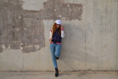 Beyzbol şapkası, ceketi ve eldiveni olan, omuzunda beyzbol sopasıyla gri çimento arka planda dinlenen genç ve güzel kızıl saçlı bir kadın. Spor ve eğlence konsepti.