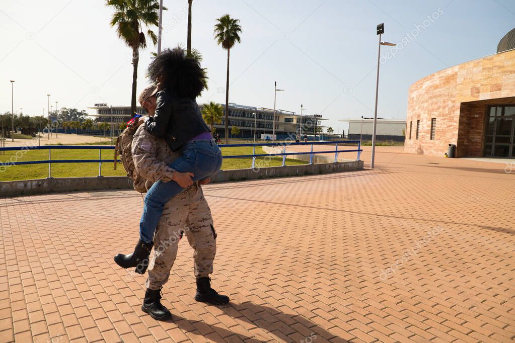 An African-American woman with an American flag jumps on her soldier boyfriend who has come home from the war. They are going to melt in a loving embrace. Concept war, soldier, army, emotion.