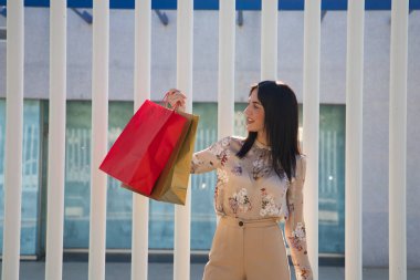 Mutluluk konsepti, tüketim, satış ve insanlar - Alışveriş torbaları tutan, gülümseyen ve alışveriş merkezinin arka planında alışveriş torbalarıyla.