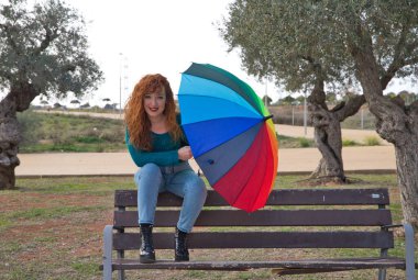 Genç ve güzel kızıl saçlı kadın birçok zeytin ağacının olduğu bir parkta bir bankta oturuyor. Gri günü aydınlatmak için bir renk şemsiyesi var. Moda ve güzellik konsepti