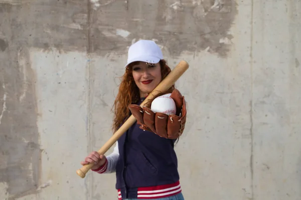 野球帽の若い美しい赤髪の女性 グレーセメントの背景に彼女の肩の上に休んで野球バットとジャケットと手袋 スポーツとレクリエーションの概念 — ストック写真