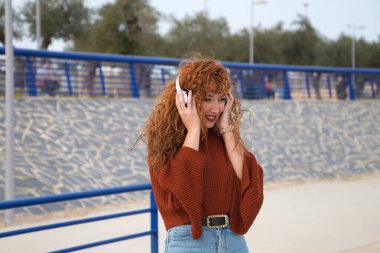 Genç ve güzel kızıl saçlı kadın büyük bir gölün olduğu bir parkta beyaz kulaklıklı müzik dinliyor. Kadın çok mutlu. İfadeler ve mutluluk, müzik ve dans.