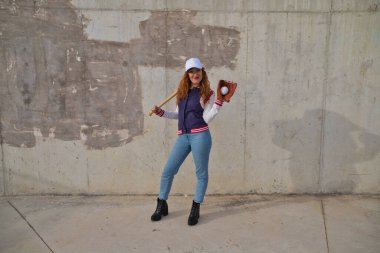 Beyzbol şapkası, ceketi ve eldiveni olan, omuzunda beyzbol sopasıyla gri çimento arka planda dinlenen genç ve güzel kızıl saçlı bir kadın. Spor ve eğlence konsepti.