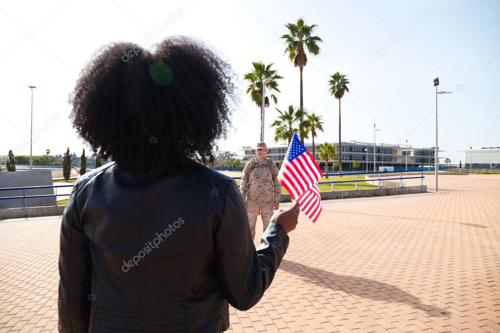 Young Afro-American woman holds two American flags in her hand while waiting for her boyfriend who has come home from the war. An American soldier can be seen in the background. Concept of war.