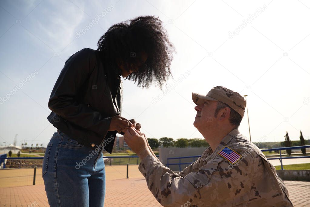 American soldier on his knees asking for marriage by holding the hands of his partner an Afro-American woman and giving an engagement ring. Concept patriotism, war, soldier.