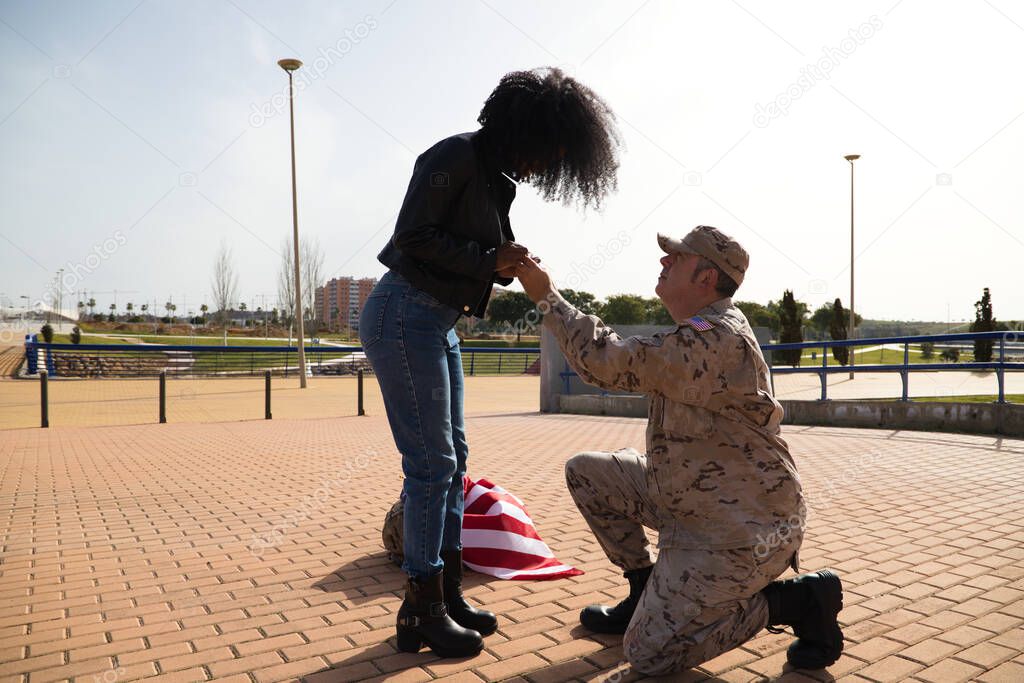 American soldier on his knees asking for marriage by holding the hands of his partner an Afro-American woman and giving an engagement ring. Concept patriotism, war, soldier.