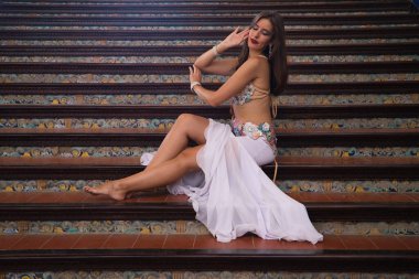 Genç ve güzel dansçı bir merdivende oturmuş dans mankeni pozları veriyor. Dansçı kırmızı bir elbise giyiyor. Göbek dansı konsepti.